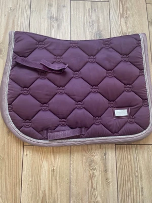 Lila schabrak från Equestrian stockholm - Säljer ett lila schabrak från Equestrian Stockholm med quiltat mönster och ljus kant. Schabraket har broderade detaljer och praktiska remmar för fastsättning. Perfekt för dig som rider och vill ha stil på utrustningen.