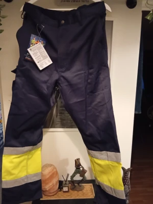 Portwest varselbyxor med reflex - Mörkblå arbetsbyxor från Portwest med gula varselpartier och breda reflexband nedtill på benen. Byxorna har klassisk rak passform och är tillverkade i slitstarkt material, perfekta för dig som vill synas extra mycket.