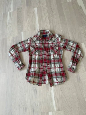 Rutig skjorta från Ralph Lauren - Säljer en rutig skjorta flanell från Ralph Lauren i rött, grönt och beige. Skjortan har långa ärmar, knappar framtill och två bröstfickor. Materialet är en mix av silke, bomull och ull för en mjuk och lyxig känsla. Slim fit-modell som sitter snyggt.