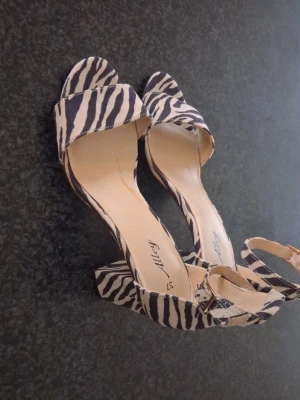 Zebramönstrade sandaler med klack stoelek 37 - Säljer ett par trendiga sandaler från Alley med zebramönster i svart och beige. Sandalerna har öppen tå, bred rem framtill och justerbart spänne vid ankeln. Klacken är bred och stabil, vilket ger både stil och komfort. Perfekta för dig som vill sticka ut!