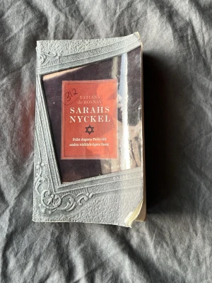 Sarahs nyckel av Tatiana de Rosnay - Romanen 'Sarahs nyckel' av Tatiana de Rosnay har ett omslag i grått och rött med en dekorativ ram och en stjärna. Boken tar dig från dagens Paris till andra världskrigets fasor. Perfekt för dig som gillar gripande och historiska berättelser.