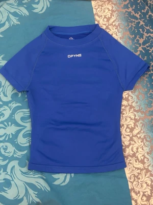 Blå sporttröja från DFYNE - Snygg blå sporttröja från DFYNE med korta ärmar och rund hals. Tillverkad i ett stretchigt funktionsmaterial som passar perfekt till träning. Logotypen är tryckt i vitt framtill och baktill vid nacken.