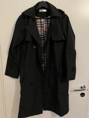Svart trenchcoat  - Klassisk svart trenchcoat med det ikoniska rutiga fodret. Jackan har dubbelknäppning, bred krage och axelklaffar. Perfekt för dig som vill ha en tidlös och stilren look. Materialet är troligtvis bomullsmix och insidan har beige, svart och rött rutmönster. Aldrig använt.