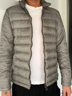 Grå peak performance pufferjacka - Snygg grå pufferjacka med ståkrage och horisontella sömmar. Jackan har dragkedja framtill och två sidofickor med dragkedja. Perfekt för kalla dagar och ger en stilren look. Materialet är mjukt och quiltat för extra värme.