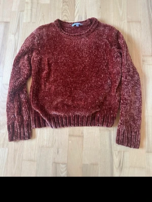  chenilletröja med rund hals - Mysig vinröd/orange brun stickad tröja i chenillematerial med rund halsringning och långa ärmar. Tröjan har ribbstickade muddar vid ärmslut och nederkant, vilket ger en skön och avslappnad look. Perfekt för att hålla värmen under kyliga dagar.