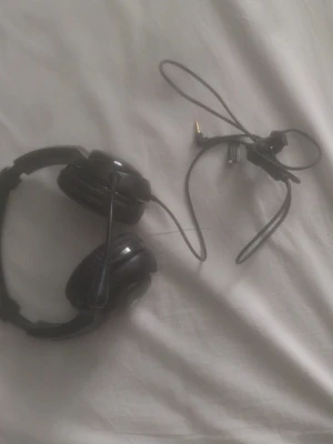 PlayStation svarta gaminghörlurar - Säljer ett par svarta gaminghörlurar från PlayStation med inbyggd mikrofon och vadderade kåpor. De har en klassisk over-ear design och ansluts med 3,5 mm kabel. Perfekta för gaming-sessioner och voice chat.