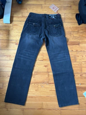 svarta True Religion jeans - Snygga svarta jeans från True Religion med raka ben och klassiska kontrastsömmar. Bakfickorna har lock och dekorativa sömmar som ger en unik look. Tillverkade i slitstarkt denim, perfekta för dig som gillar streetwear och vill sticka ut.