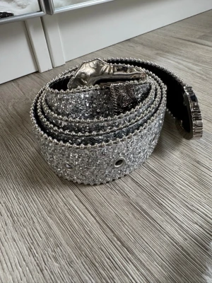 Glittrigt silverbälte med detaljer - Säljer ett brett bälte i silver med glittrig yta och dekorativa nitar längs kanterna. Spännet är stort och har snygga mönstrade detaljer i metall. Perfekt för att ge outfiten extra bling och statement. 3.8 bred 122cm lång