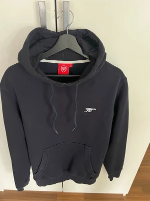 Arsenal hoodie med logga - Mörkblå hoodie från Arsenals officiella klubbshop med kanon broderad på bröstet. Klassisk känguruficka framtill och justerbar huva med snören. Perfekt för dig som vill visa ditt stöd för laget på ett stilrent sätt. StrL (ganska nära M)