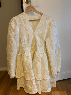 Vit omlottklänning med volang - Supersöt vit kortklänning med omlott och volangdetaljer. Klänningen har långärmade puffärmar och ett strukturerat tyg med diskret mönster. Perfekt för sommarens alla tillfällen och ger en romantisk vibe. Använd 1 gång. Inga defekter