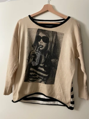 Beige tröja med tryck och strass - Trendig beige tröja med svartvitt fototryck framtill och strassdetaljer på motivet. Tröjan har svarta kanter vid hals och nederkant samt randigt tyg i svart och vitt på insidan och baktill. Långärmad och mjuk, perfekt för en cool vardagslook.