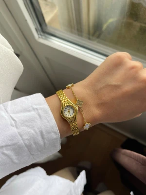 Guldfärgad klocka med guldfärgade fyrklöver armband  - Stilren guldfärgad klocka med klassiskt länkarmband och rund vit urtavla. Klockan har svarta siffror och visare, vilket gör den enkel att läsa av. Perfekt accessoar för dig som gillar en elegant och tidlös look. Bandet är legering på klockan, själva klockan är stål, 19cm lätt att justera med en nål. Armbandet är rostfritt stål och guldplöterad. 18+5.