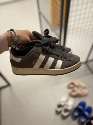 Adidas Campus grå sneakers - Säljer ett par grå Adidas Campus sneakers med klassiska vita ränder på sidorna och vit häl. Skorna har breda svarta skosnören och en beige sula. Ovandelen är i mocka och tungan har Adidas-loggan i vitt. Perfekta för dig som gillar retrostil.