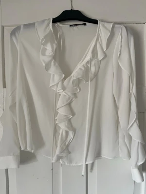 Vit volangblus från Zara - Säljer en vit blus från Zara med långa ärmar och snygga volangdetaljer längs urringningen. Blusen har en luftig passform och tunna knytband framtill. Perfekt för dig som gillar en romantisk och trendig stil.