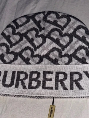Grå Burberry mössa med hjärtmönster - Säljer en snygg grå mössa från Burberry med mörkgrått hjärtmönster och stor logotyp framtill. Mössan är stickad i mjukt material och har en bred kant med BURBERRY-text. Perfekt accessoar för att lyfta din outfit.