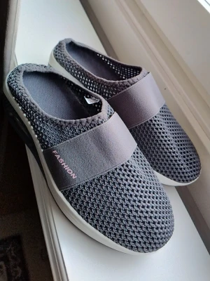 Mörkgrå mesh-tofflor med resårband - Säljer ett par mörkgrå tofflor med ventilerande meshmaterial och breda elastiska band över foten. Sulan är vit och ger en sportig look. Perfekta för dig som vill ha något lätt och bekvämt att glida i hemma eller snabbt ut. Märkta med 'FASHION' på bandet.