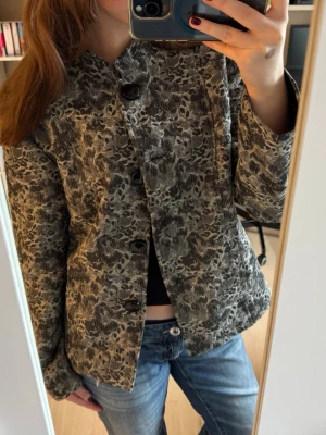 Gråmönstrad jacka med stora knappar - Snygg jacka från Cappuccini i grått och svart med blommigt mönster. Rak passform, stora svarta knappar framtill och lång ärm. Materialet känns lite tjockare och ger en exklusiv känsla. Perfekt för dig som vill sticka ut med en unik jacka.