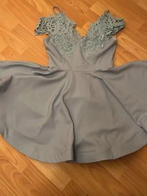 Ljusblå spetsklänning från Ginger Fizz - Supersöt ljusblå klänning från Ginger Fizz med v-ringning och detaljerad spets upptill. Kjolen är utsvängd och klänningen har smala axelband. Perfekt för dig som gillar romantiska detaljer och en feminin look.