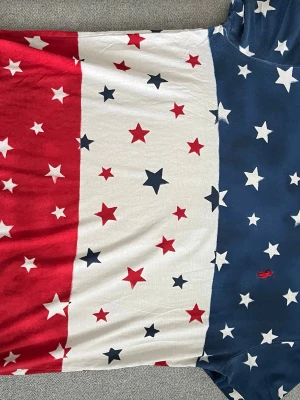 Stjärnmönstrad t-shirt från Polo Ralph Lauren - Snygg t-shirt från Polo Ralph Lauren med stjärnmönster i blått, vitt och rött. Klassisk rund hals och korta ärmar. Ikoniska Polo-loggan broderad på bröstet. Perfekt för dig som vill sticka ut med en färgstark och unik look.