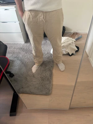 Beige linnebyxor med dragsko - Säljer ett par beige linnebyxor med avslappnad passform och dragsko i midjan. Byxorna har sidofickor och är perfekta för en chill och stilren look. Materialet är lätt och luftigt, vilket gör dem sköna att bära under varma dagar.