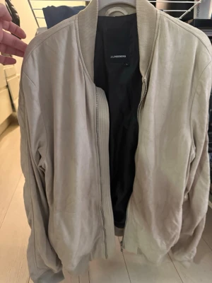 Beige bomberjacka från J.Lindeberg - Stilren beige bomberjacka från J.Lindeberg med mjuk mockaliknande yta. Jackan har ribbade muddar vid krage, ärmslut och nederkant samt dragkedja framtill. Perfekt för dig som vill ha en clean och modern look.