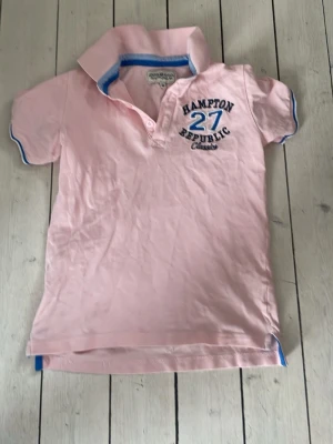 Rosa piké från Hampton Republic - Snygg rosa pikétröja från Hampton Republic med blå och vita detaljer på krage och ärmslut. Tryck med text och siffror på bröstet. Klassisk pikékrage och korta ärmar. Perfekt till en sommar stämning!