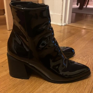 Svartlackade boots med mellanhög klack - Säljer ett par svarta stövletter i glansigt lackmaterial med spetsig tå och bred klack. Skorna har dragkedja på insidan och en cool, modern look som passar till många outfits. 