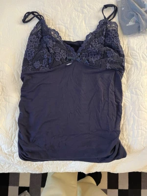 Mörkblå spetsnattlinne med rosett - Sött nattlinne i mörkblått med tunna axelband och spetsdetaljer över bysten. Dekorerad med en liten rosett framtill och blommig spets. Mjuk och följsam känsla, perfekt för mysiga kvällar.