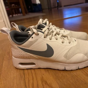 Nike AirMax vit sneakers med gråa detaljer - Snygga vita Nike sneakers med grå swoosh på sidan. Skorna har en luftig mesh-överdel, vita snören och en vadderad insida i mörkgrått. Sulan är tjock och har synlig Air-enhet i hälen för extra komfort vid gång. Använda måttligt.