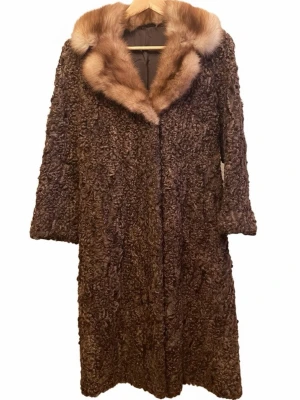 Brun pälsrock med krage i räv - Lång, mörkbrun pälsrock med bred krage i ljusbrun räv. Kappan har en klassisk rak passform och är tillverkad i lockig fårpäls med lyxig känsla. Perfekt för dig som vill ha en statement piece med vintagevibbar och extra värme under kalla dagar.