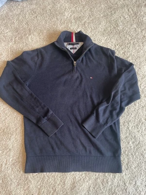 Mörkblå halvzip från Tommy Hilfiger - Snygg mörkblå stickad tröja från Tommy Hilfiger med halv dragkedja och liten broderad logga på bröstet. Tröjan har ribbade muddar och krage med röd och vit detalj på insidan. Perfekt till lager på lager och enkel att matcha.