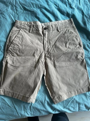 Beige bomullsshorts med fickor - Snygga beige shorts i bomull med normal passform. Klassisk design med bälteshällor, knapp och dragkedja framtill samt två sidofickor och två bakfickor. Perfekta för varma dagar och enkel att matcha med olika stilar.