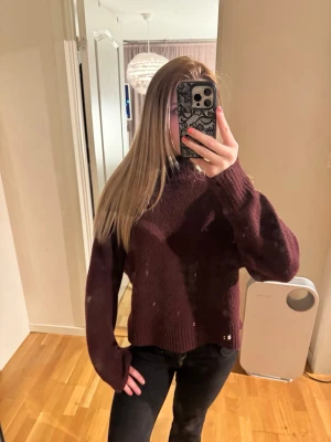 Mörkröd stickad polotröja - Säljer en mörkröd stickad polotröja med avslappnad passform och långa ärmar. Tröjan har ribbade muddar vid ärmslut och nederkant samt en hög hals som ger extra mysfaktor. Perfekt för kalla dagar och enkel att styla med jeans eller kjol.