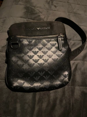 Svart axelväska Emporio Armani - Snygg svart axelväska från Emporio Armani med präglat logomönster över hela väskan. Väskan har justerbar axelrem, dragkedja upptill och detaljer i metall. Materialet är konstläder med textilparti upptill där loggan är tryckt i silver. Nypris 1800