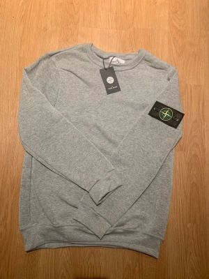 Grå sweatshirt från Stone Island - Säljer en stilren grå sweatshirt från Stone Island med klassisk rund hals och ribbade muddar. Tröjan har den ikoniska Stone Island-patchen på vänster ärm och är tillverkad i mjukt bomullsmaterial. Perfekt för dig som gillar streetwear och vill ha en clean look.