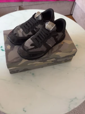 Valentino Garavani camo sneakers - Snygga sneakers från Valentino Garavani med svart och grått camomönster. Skorna har en mix av mocka och mesh, svarta snören och en klassisk låg siluett. Perfekta för dig som vill ha en unik och trendig look.