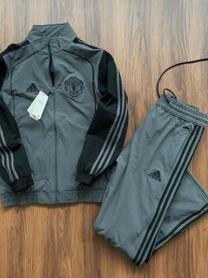 Grå Manchester United träningsset Adidas - Snyggt grått träningsset från Adidas med Manchester United-logga på jackan. Setet består av en zip-jacka och matchande byxor, båda med svarta detaljer och klassiska Adidas-ränder längs ärmar och ben. Perfekt för träning eller chill. Materialet är lätt och sportigt.