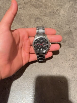 Silver färgad klocka från Aviator  - Stilren analog klocka från Aviator med svart urtavla och detaljer i rött och vitt. Klockan har en robust länk i silverfärgat metall och flera små urtavlor för extra funktioner. 