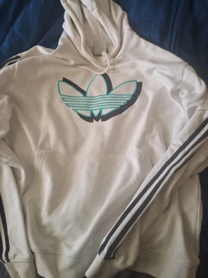Vit Adidas hoodie med turkost tryck - Säljer en vit hoodie från Adidas med stor broderad turkos och svart Trefoil-logga på bröstet. Klassiska svarta ränder längs ärmarna och känguruficka framtill. Mjuk bomullsblandning och justerbar huva med snören. Perfekt för en sportig och avslappnad stil.