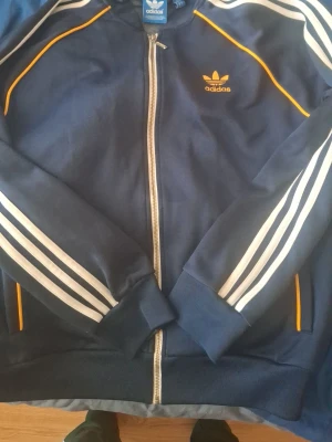 Blå Adidas zip-tröja med ränder - Som ny ! Snygg blå zip-tröja från Adidas med klassiska vita ränder längs ärmarna och gula detaljer. Tröjan har dragkedja framtill, ribbade muddar och Adidas logga på bröstet. Perfekt för en sportig look.