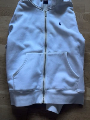 Vit hoodie från Polo Ralph Lauren - Säljer en klassisk vit hoodie från Polo Ralph Lauren med dragkedja framtill och känguruficka. Ikoniska blå loggan broderad på bröstet. Hoodien har huva och är gjord i mjukt bomullsmaterial, perfekt för en clean och stilren look.