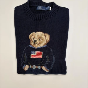 Mörkblå stickad tröja Polo Bear - Mörkblå stickad tröja från Polo Ralph Lauren med ikoniskt Polo Bear-motiv framtill. Björnen har en tröja med amerikanska flaggan och jeans, vilket ger en cool och klassisk look. Rund halsringning och ribbade muddar. Perfekt för dig som gillar preppy stil.