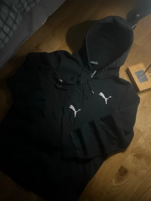 Svart hoodie från Puma med logga - Säljer en svart hoodie från Puma med klassisk vit logga på bröstet. Hoodien har huva med dragsko och är tillverkad i mjukt material som är perfekt för en avslappnad och sportig look. Passar dig som gillar streetwear och enkel stil och T-shirt.
