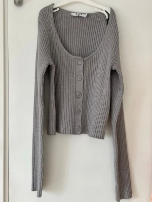 Grå kofta från NA-KD - Säljer en grå ribbad kofta från NA-KD med djup rundad halsringning och knappar framtill. Koftan har långa ärmar och är tillverkad i ett mjukt, stretchigt material som sitter snyggt på kroppen. Perfekt att matcha med jeans eller kjol.