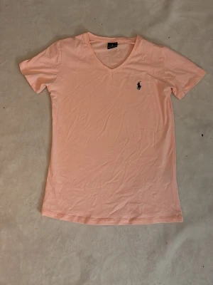 Ljusrosa t-shirt från Polo Ralph Lauren - Ljusrosa t-shirt från Polo Ralph Lauren med v-ringning och klassisk broderad logga på bröstet. T-shirten är i mjuk bomull och har en slim fit passform. Perfekt för en avslappnad och stilren look.