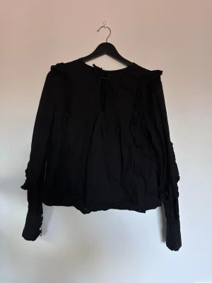 Svart blus med volangdetaljer - Svart blus från bikbok med volangdetaljer längs axlar och ärmar. Blusen har långärmad design med knappar vid ärmslut och knytning i halsen. Materialet är lätt och flowy, perfekt för att skapa en cool look.
