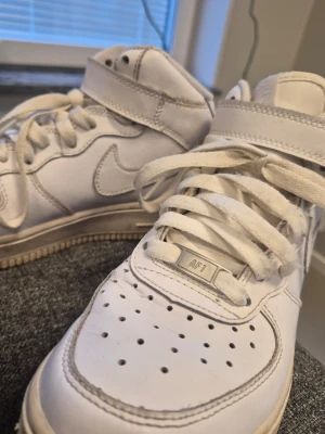 Nike Air Force 1 vita high sneakers 36 - Nike Air Force 1 high sneakers i helvitt läder med klassisk swoosh på sidan och perforerad tå. Skorna har högt skaft med kardborreband och vita skosnören. Perfekt för dig som gillar ikoniska och stilrena sneakers.