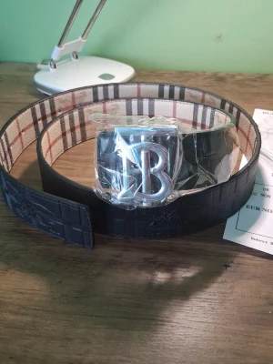 Burberry bälte med silver B-spänne - Snyggt Burberry bälte i svart skinn med klassiskt rutigt mönster på insidan och ett stort silverfärgat B-spänne framtill. Kommer med originalpåse och box. Perfekt accessoar för att lyfta din outfit. 110cm!