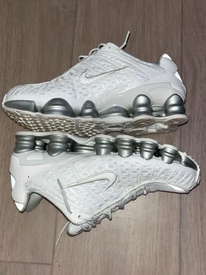 Nike Shox vita sneakers - Nike Shox sneakers i vitt med silverfärgade fjädrande sulor. Ovandelen är i mesh och syntetmaterial med tydliga detaljer och snörning. Ikonisk Swoosh-logga på sidan och sportig design som sticker ut.