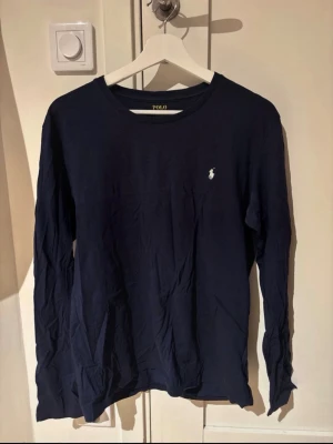 Marinblå långärmad topp Polo Ralph Lauren - Snygg marinblå långärmad topp från Polo Ralph Lauren med klassisk rund halsringning och den ikoniska vita logobrodyr på bröstet. Tillverkad i mjuk bomull som känns skön mot huden. 